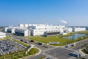 Aerial view of a large industrial factory with parking lots and smoke emissions under a clear blue sky.